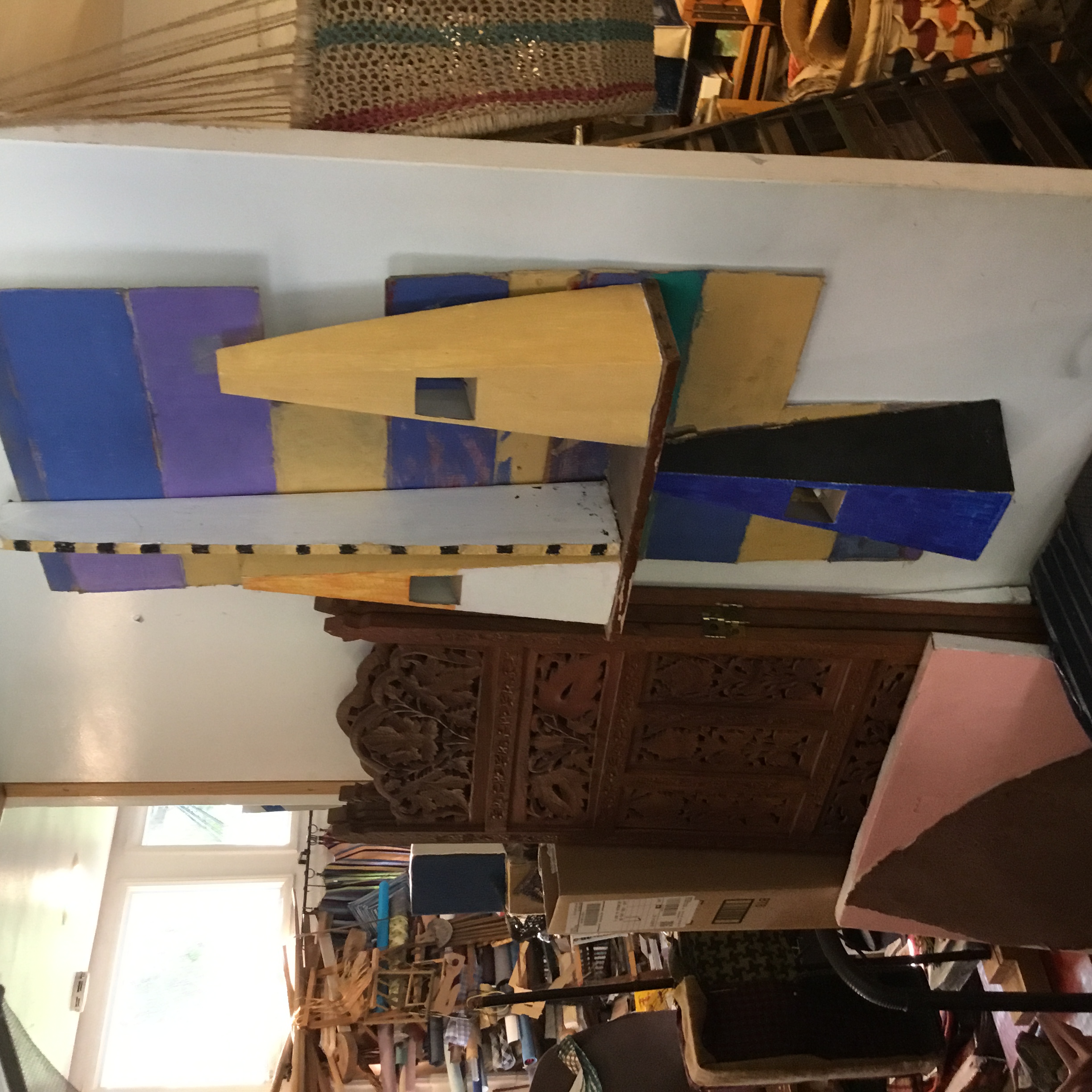 This work is on a flattened box. The three main structures, orange and white, yellow, and blue, are cardboard box inserts. The vertical piece that is white, yellow, and black is a piece of dry wall. These parts have all been painted with water pastels and acrylic paint. The shelf that holds two of the box inserts is a piece of wood.