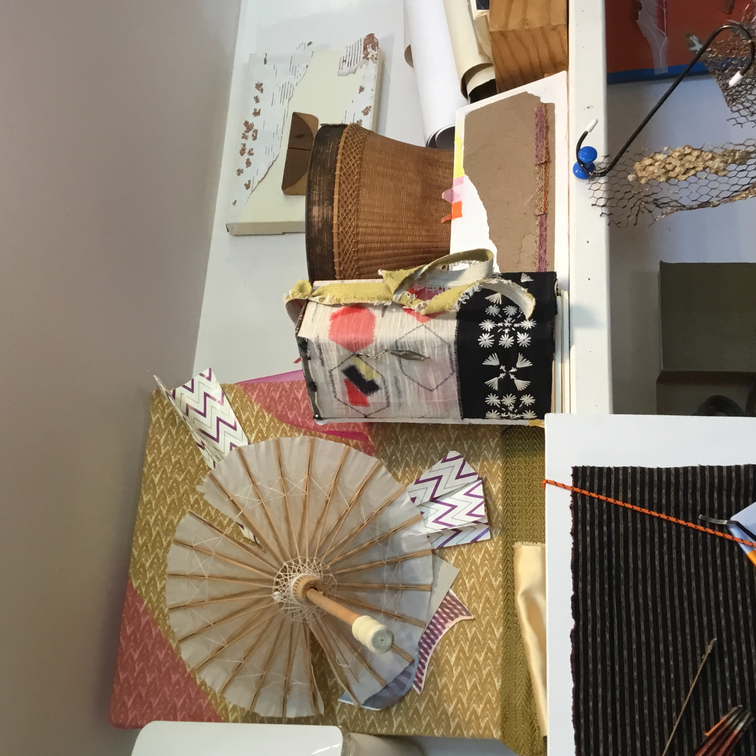 This picture contains two works. The piece of the left is a cloth covered canvas with kite paper from Germany and an open umbrella. The piece on the right is a box covered in a Japanese fabric with a hexagon design. The black and white patterned cloth below it is from Thailand. A shade of yellow fabric was originally used to tie the inside of a package together. There is a piece of metal hanging down with a bead and flower design.