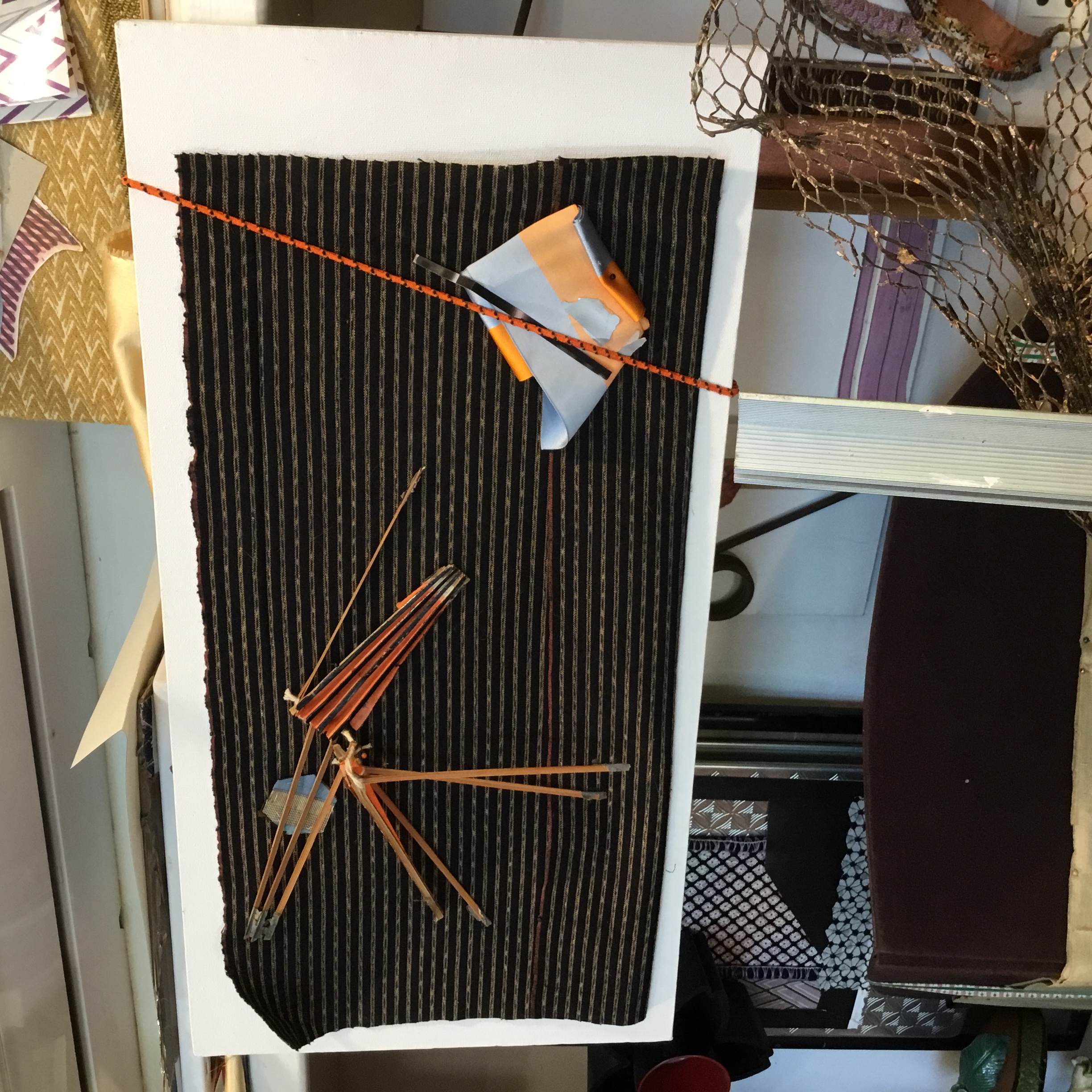 This work is Japanese indigo fabric that is attached to a canvas. On the left are detached umbrella spokes. The right corner contains a folded, blue and orange ribbon that is attached with a bungie cord.