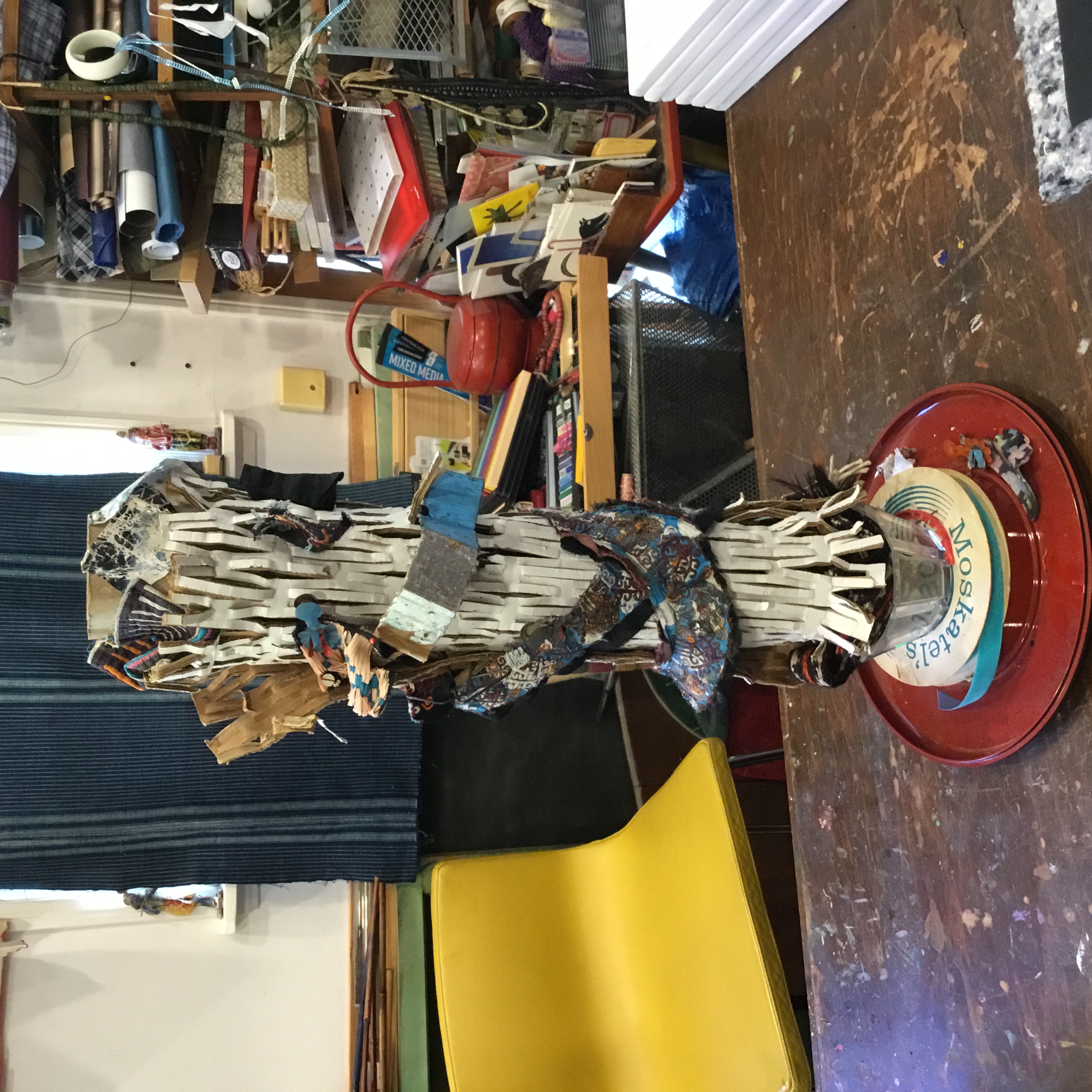 From bottom to top: This tower has Japanese lacquerware as the base. A ribbon spool is beneath a transparent, plastic bowl. Inside the bowel, a camel strap is inserted. Above the bowl is a Uzbek blue and white collar and a piece of mola. The white structure is packing material.